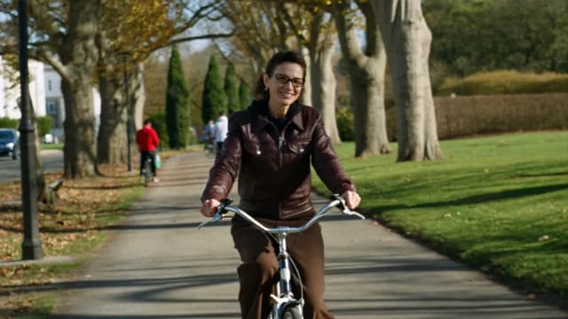 A woman cycling toward the camera along a tree-lined park path
