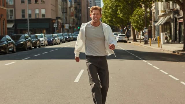 A man in a white jacket walking toward the camera down a sunlit avenue